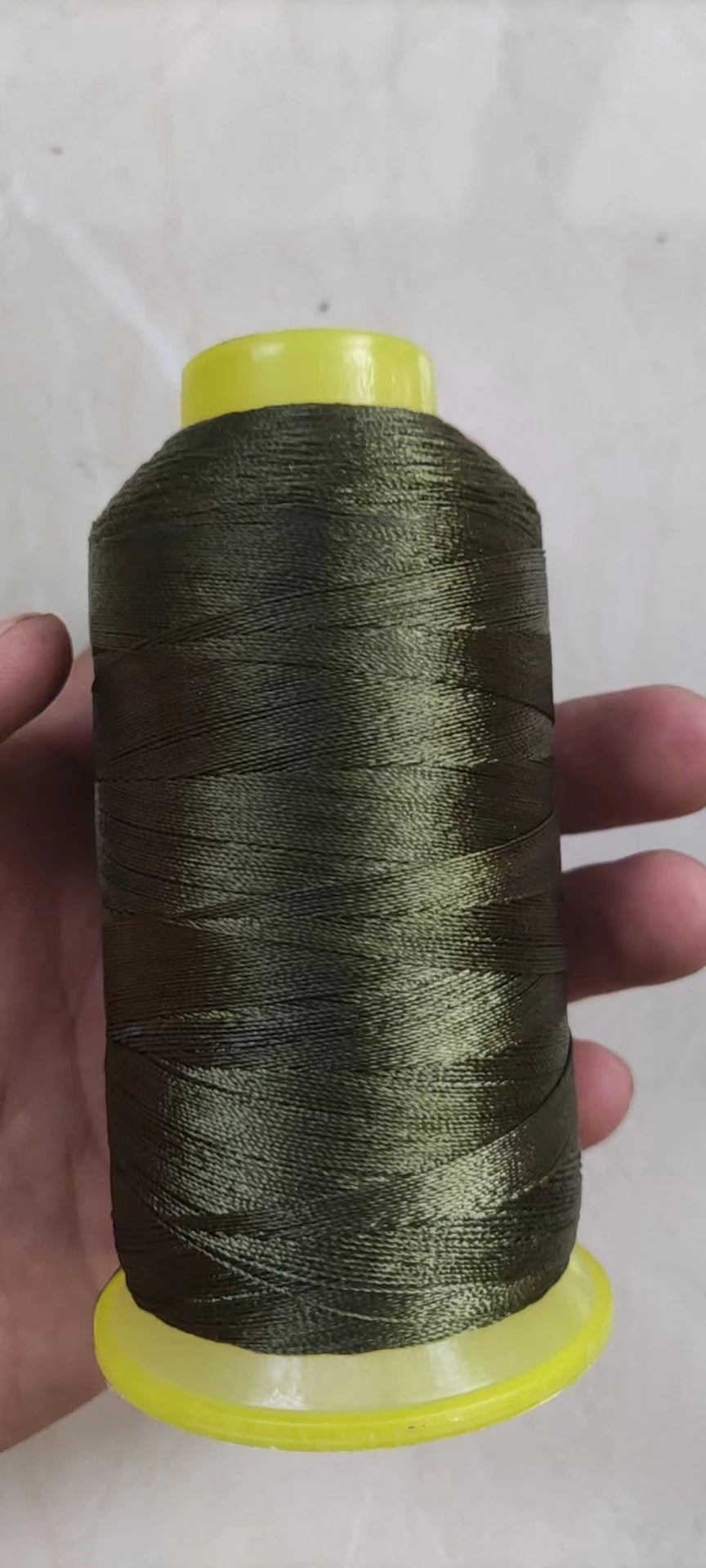 A spool of dark olive green high tenacity polyester sewing thread with a yellow plastic core.