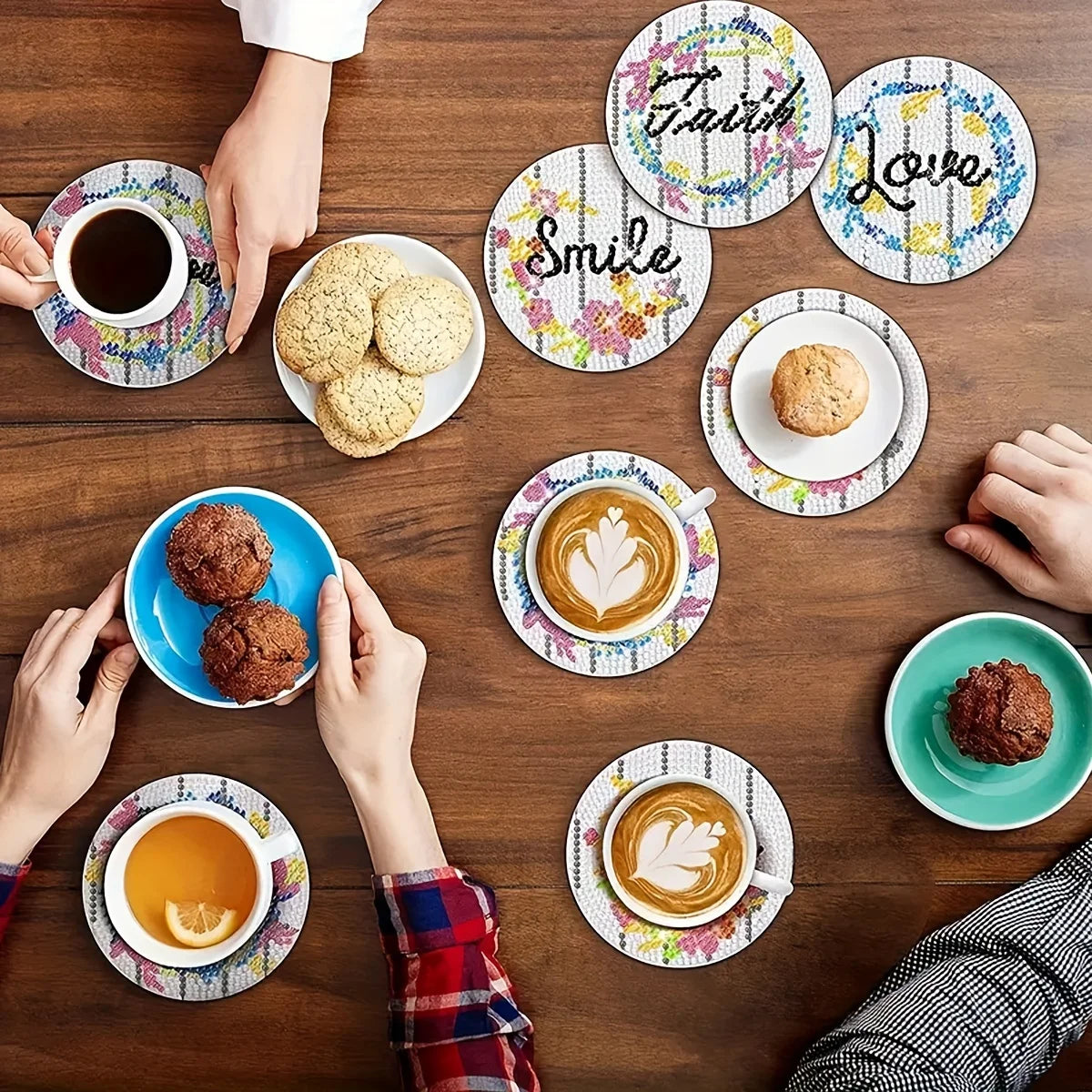 A flat lay of various coffee cups, cookies, and a diamond painting coaster set on a wooden table.