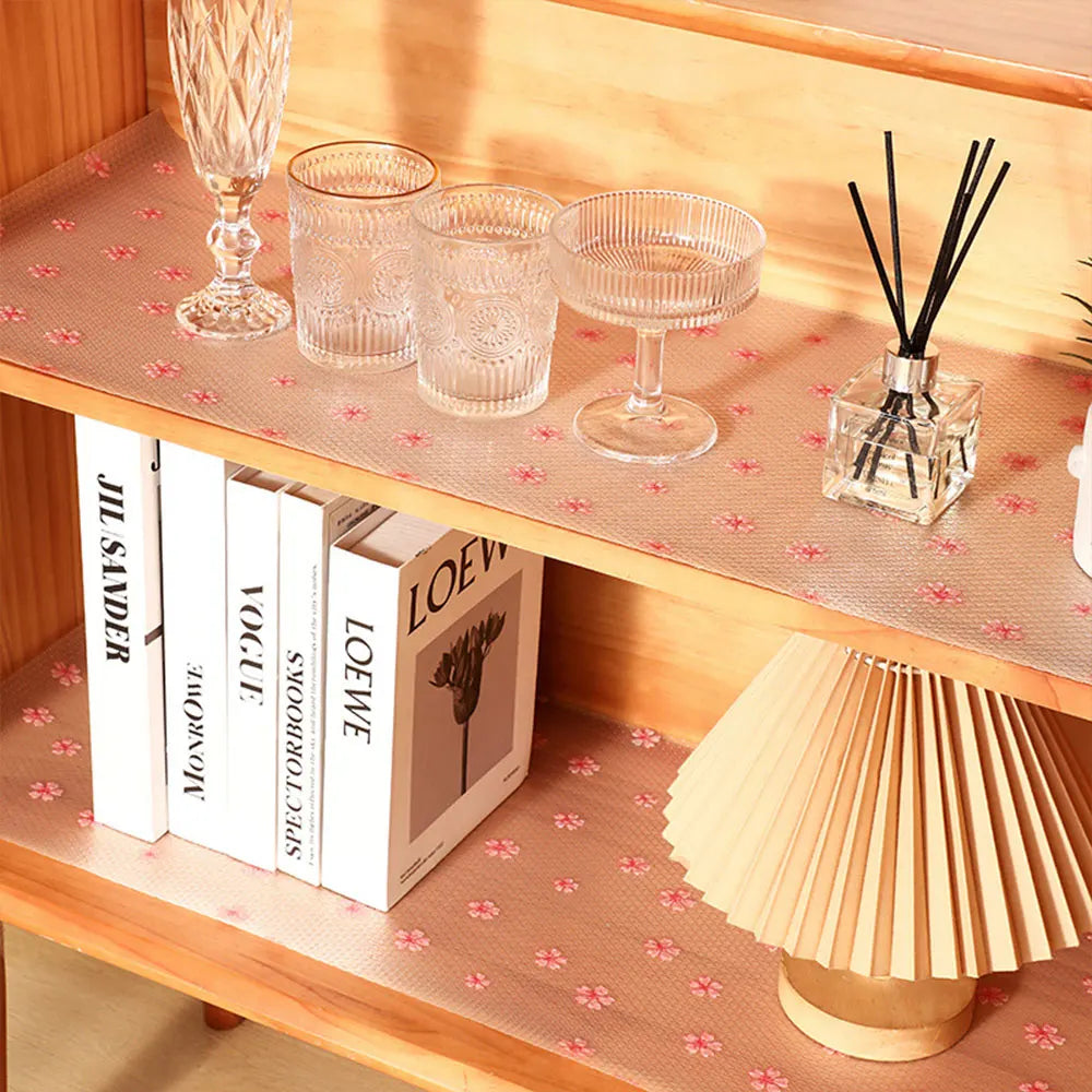 A wooden shelf displays glassware, books, and a decorative lamp shade on a pink patterned surface.