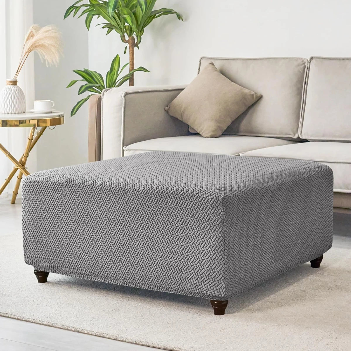 A gray stretch jacquard ottoman cover placed on a light rug next to a beige sofa and potted plant.