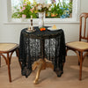 A dark lace tablecloth is set on a wooden table with a vase and small food items in front of a bright window.