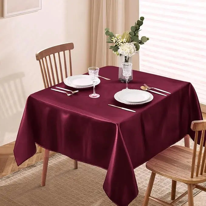 A deep burgundy satin tablecloth is set for dining with white plates and silverware, decorated with a floral arrangement.