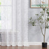 A white vintage floral lace curtain panel hangs in front of a window with a framed nature painting and a potted plant.