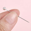 A hand holds a silver sewing pin with a pearl head against a light pink background.