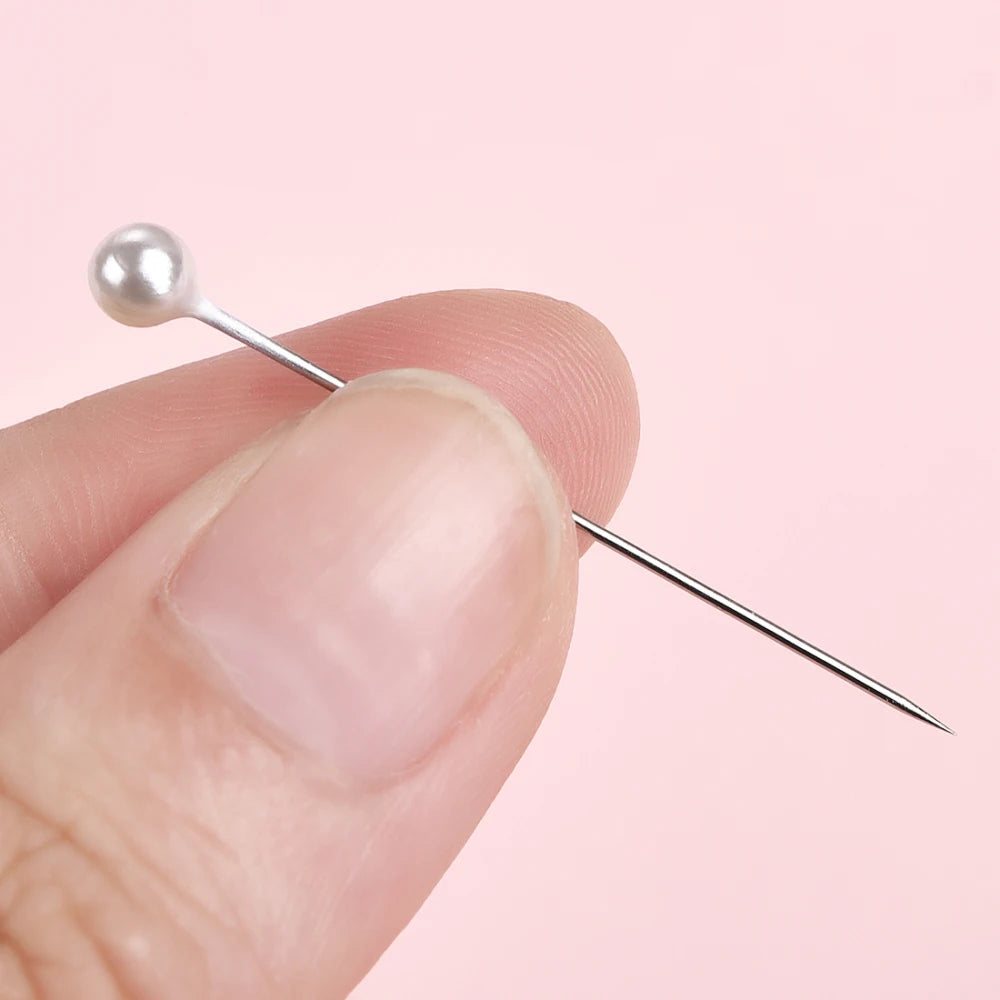 A hand holds a silver sewing pin with a pearl head against a light pink background.