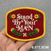 A maroon embroidered patch with the text Stand By Your MAN and floral accents on a yellow border.