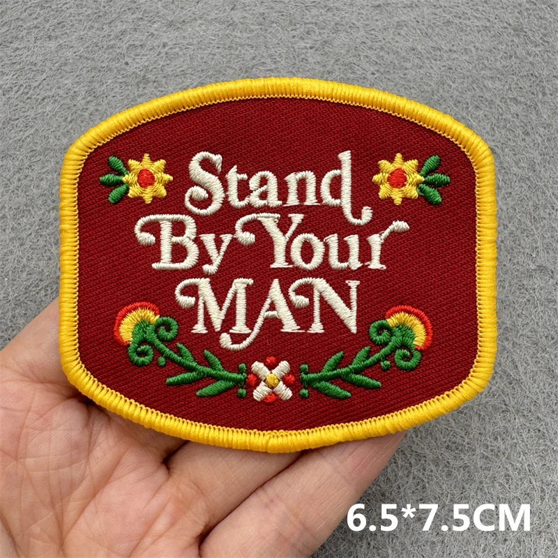 A maroon embroidered patch with the text Stand By Your MAN and floral accents on a yellow border.