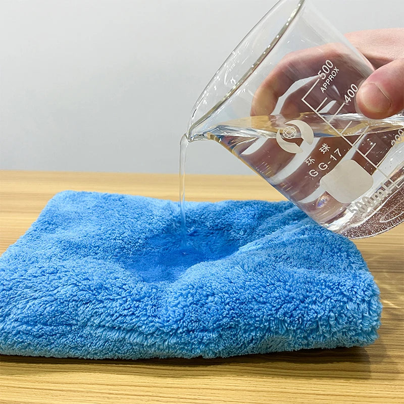 A hand pours liquid into a clear glass measuring cup, dripping onto a bright blue coral fleece microfiber cleaning cloth.