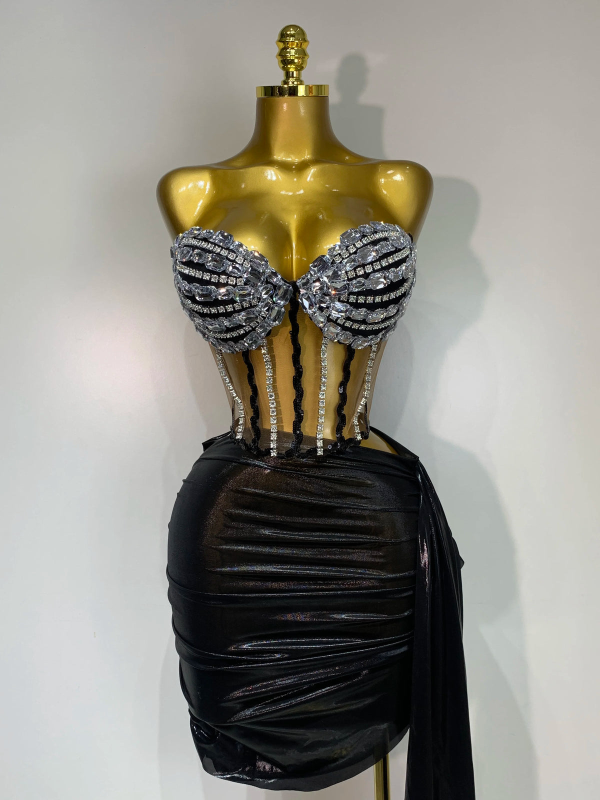 A gold mannequin displays a sparkly diamond two-piece performance set featuring embellished cups and a black draped skirt.