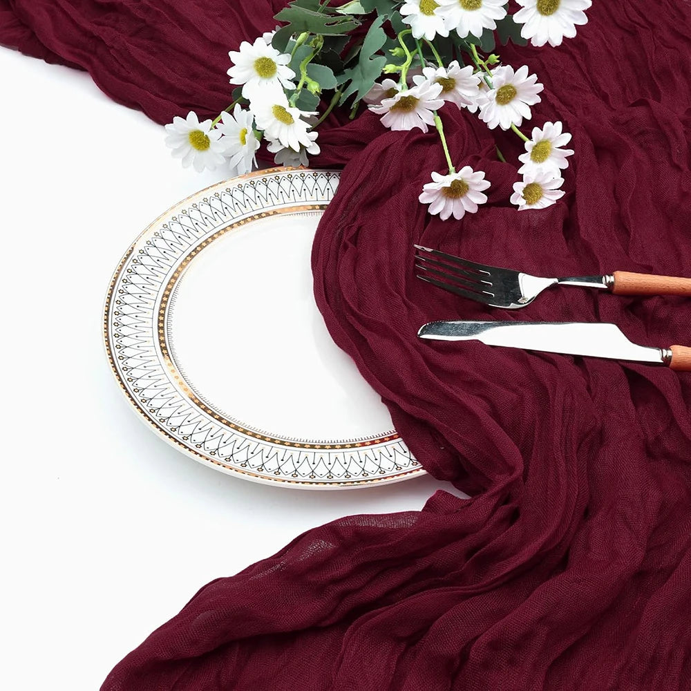 A deep burgundy cheesecloth table runner draped with white flowers, featuring a decorative plate and silverware.