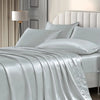 A luxurious silver satin pillowcase draped over a bed with white pillows and a bedside lamp.