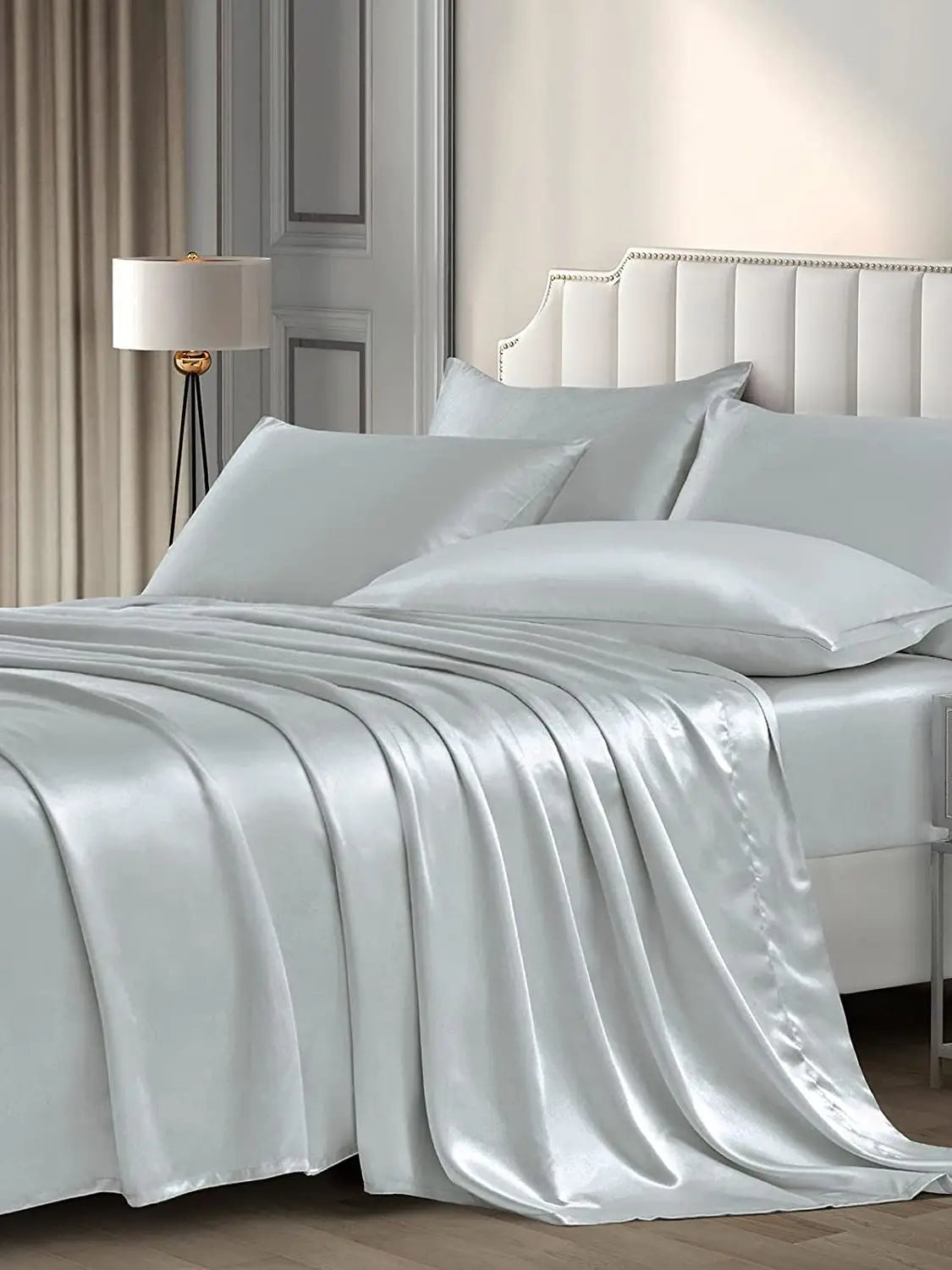 A luxurious silver satin pillowcase draped over a bed with white pillows and a bedside lamp.