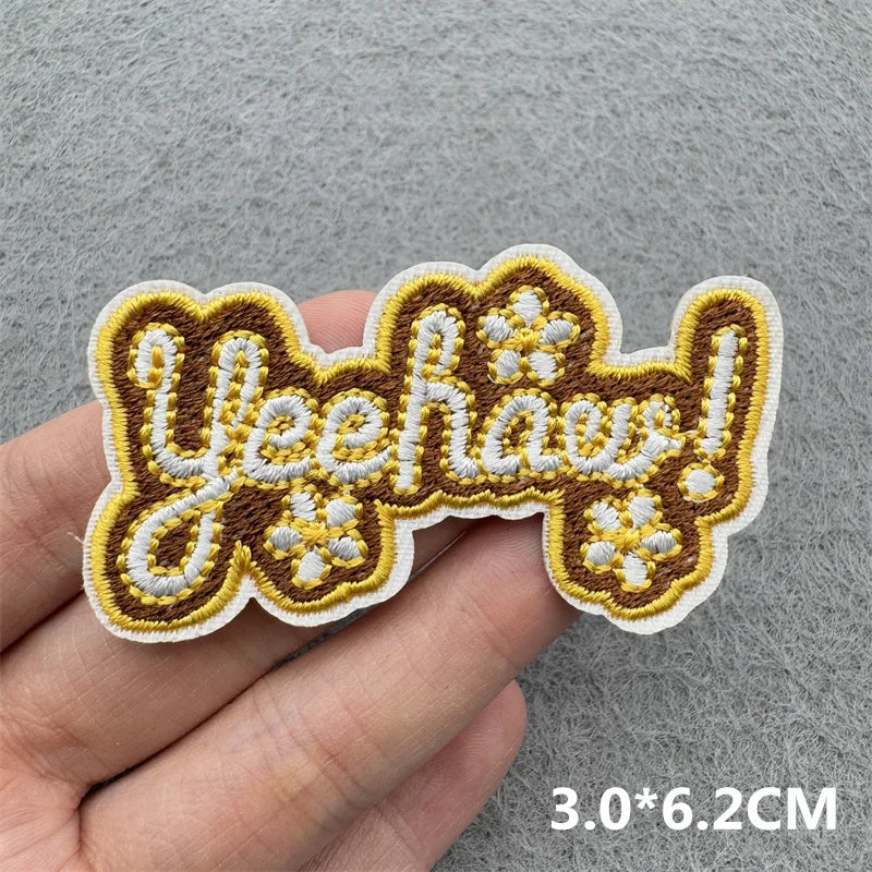 A hand holds a yellow and brown embroidered iron-on patch with the text Yeehaaw!