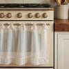 A white kitchen curtain skirt with a ruffle trim and ribbon ties hanging on a beige stove.
