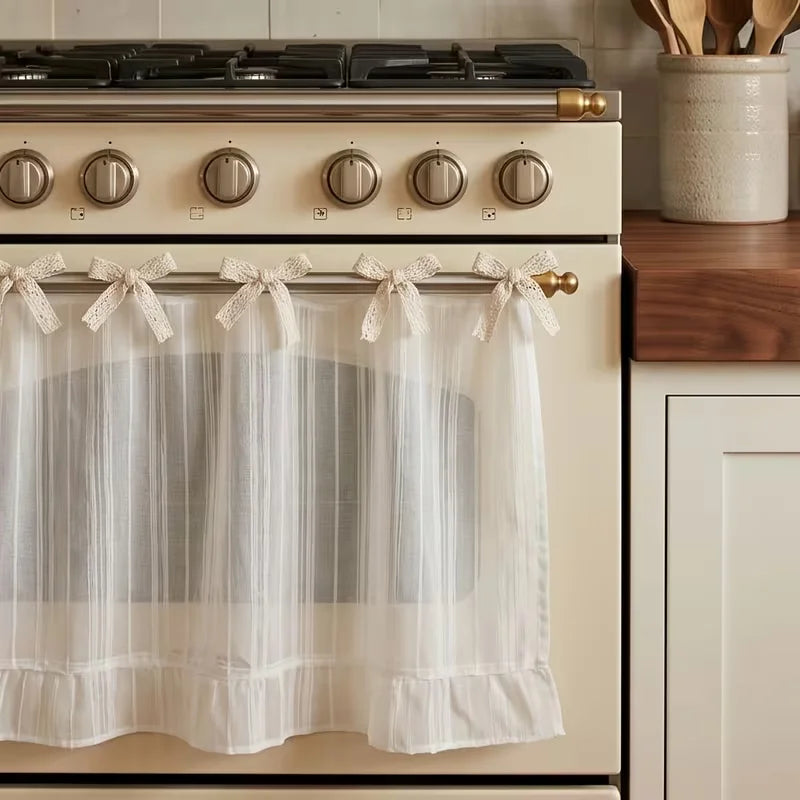 A white kitchen curtain skirt with a ruffle trim and ribbon ties hanging on a beige stove.