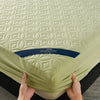 A hand is holding a light green quilted mattress protector with a blue label, showing the texture and fit.