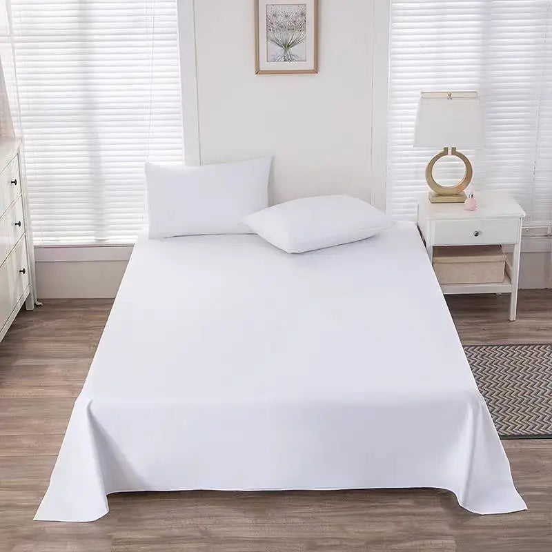 A white premium flat bed sheet is neatly laid on a bed with white pillows, set against a window with sheer white blinds.