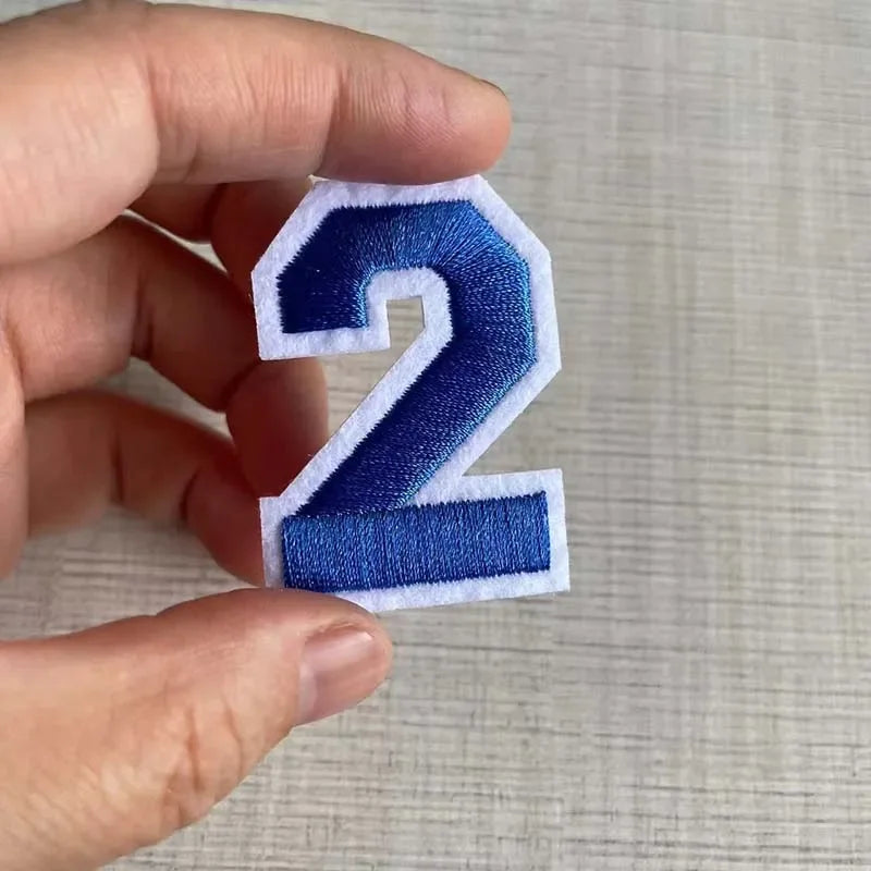 A hand holds a blue embroidered number 2 patch on a light background.