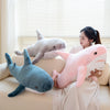 A woman is holding three large plush shark toys of gray, pink, and blue colors indoors.
