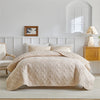 A beige quilted bedding set spread on a bed with white paneled walls and a mirror.
