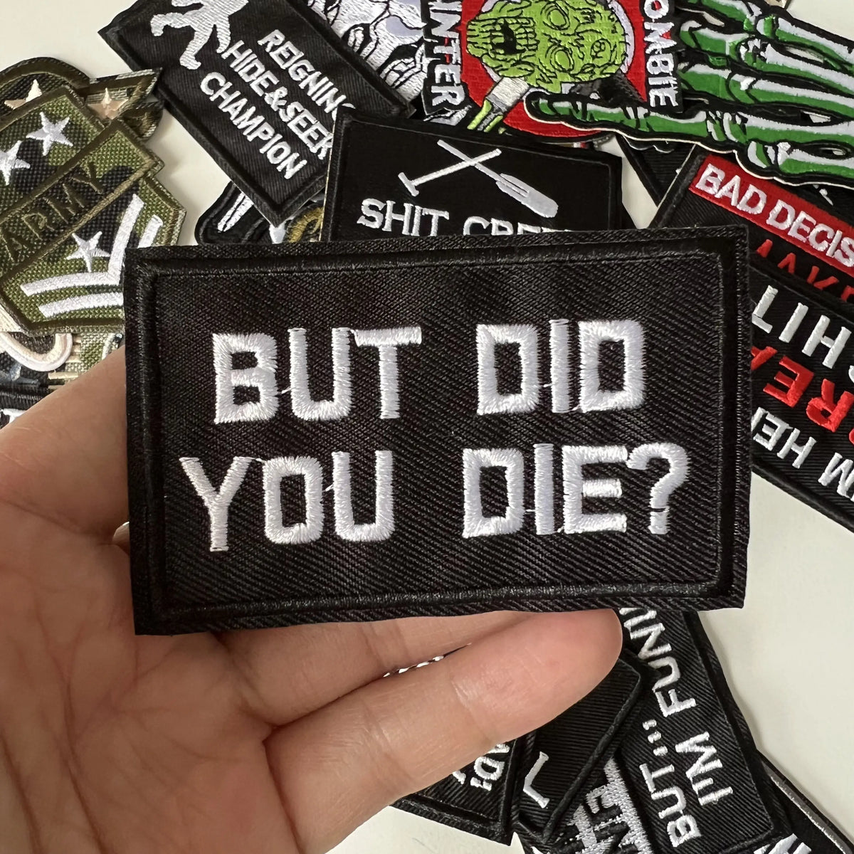 A hand holds a black iron-on slogan patch with white text that reads BUT DID YOU DIE?