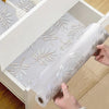 Roll of white silicone non-slip drawer liner with silver leaf pattern being pulled out of a storage box