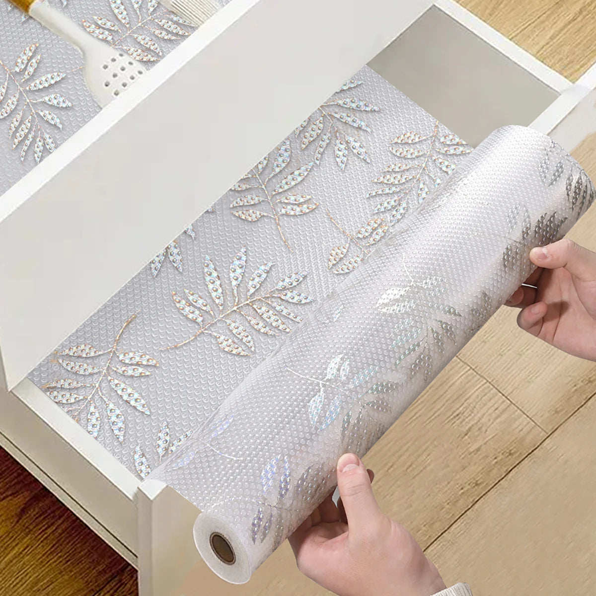 Roll of white silicone non-slip drawer liner with silver leaf pattern being pulled out of a storage box