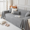 A light gray anti-scratch sofa cover draped over a modern sofa with decorative pillows and a small cup on the fabric.