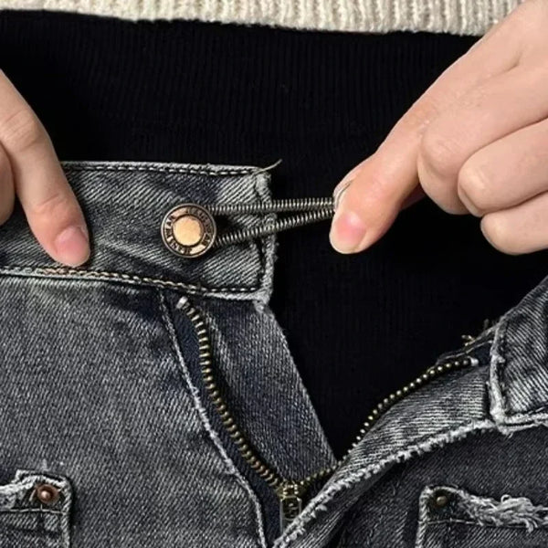A hand is using a metal button extender to adjust the waistband of dark denim jeans.