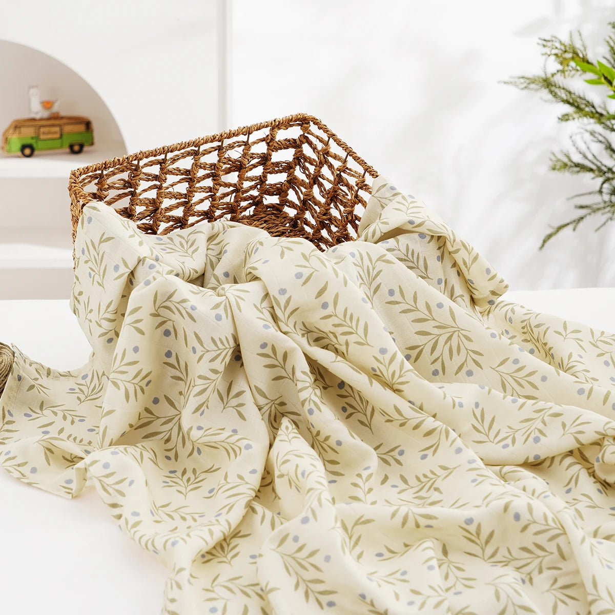 A cream-colored muslin swaddle blanket with a delicate leaf pattern is draped over a woven basket.