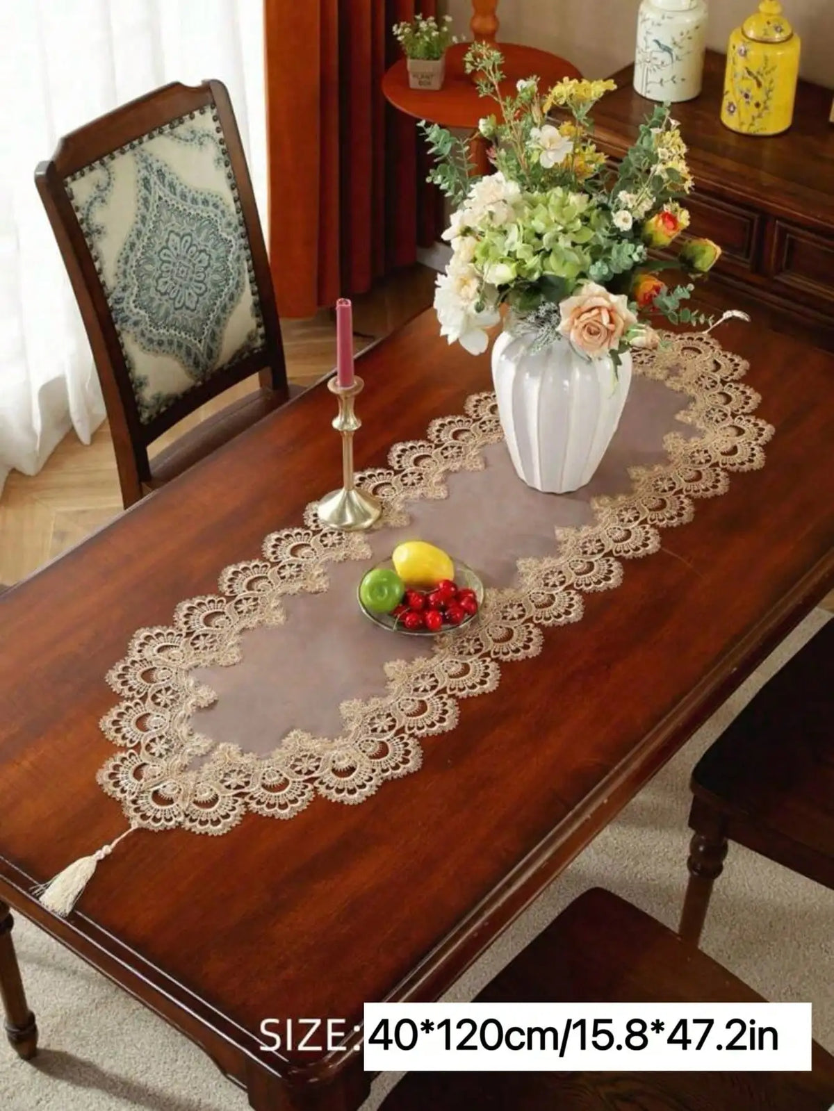 A brown wooden table is decorated with a beige embroidered mesh lace table runner featuring fresh fruit and flowers.