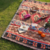 A woven blanket with geometric patterns and fringe laid on green grass, featuring a wooden guitar, orange fruit, and pink fab