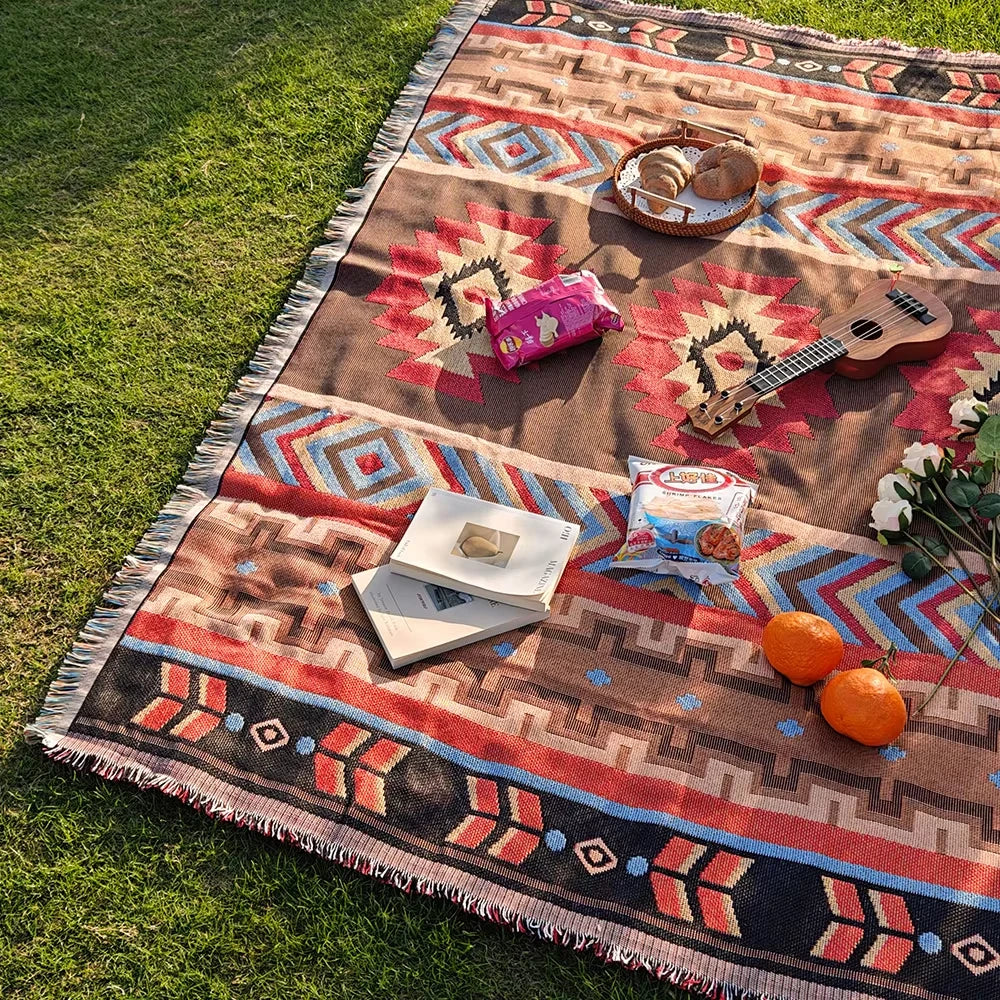 A woven blanket with geometric patterns and fringe laid on green grass, featuring a wooden guitar, orange fruit, and pink fab