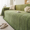 A textured olive green sofa cover featuring an elegant fishbone pattern, with a small pillow and a rolled cream throw visible