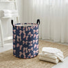 A navy blue canvas laundry basket with a pink bow pattern sits on a woven mat next to white towels.