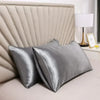 A pair of smooth, silver-gray mulberry silk pillow covers resting on a bed with a padded headboard and a white lamp.