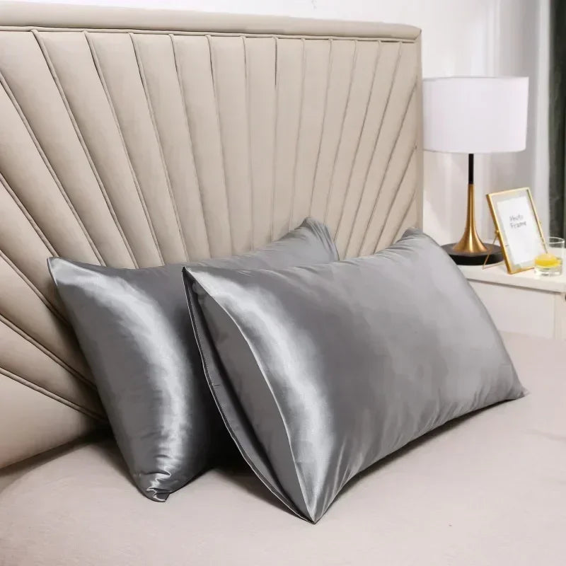 A pair of smooth, silver-gray mulberry silk pillow covers resting on a bed with a padded headboard and a white lamp.