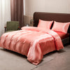 A pink satin luxury bedding set is displayed on a bed with a dark brown headboard and light curtains.