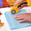 A hand holds a yellow precision acrylic quilting ruler with a dual scale and seam rail placed on a blue grid paper.