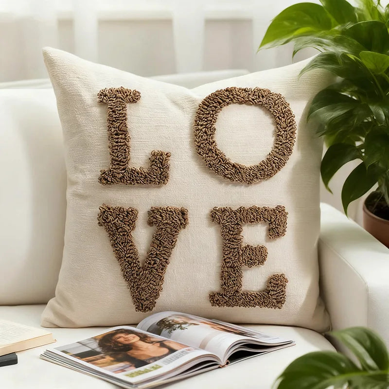 A beige knit pillow cover with the word LOVE embroidered in brown yarn rests on a white sofa.