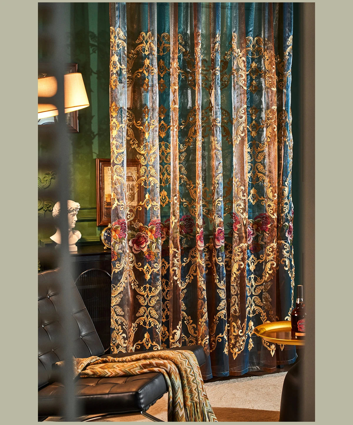 Long, sheer teal curtains with gold floral embroidery hang in a room next to a dark leather chair.