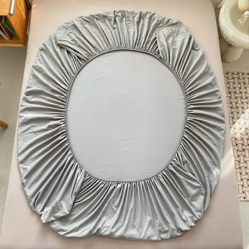 A round fitted bed sheet in silver gray fabric is displayed on a light pink surface.