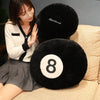 A woman is sitting next to a black fuzzy 8 ball plush pillow on a beige sofa.