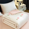 A white embroidered cooling summer quilt with pink floral designs is folded on a bed, next to a bedside table.