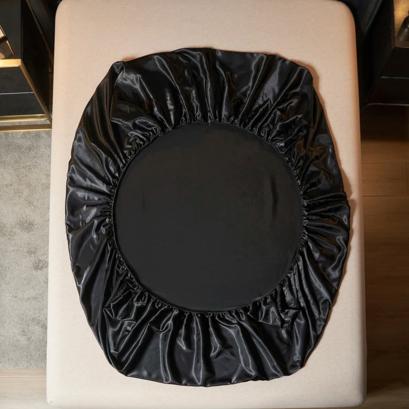 A black satin bedding set with an elastic band is displayed on a light-colored surface.