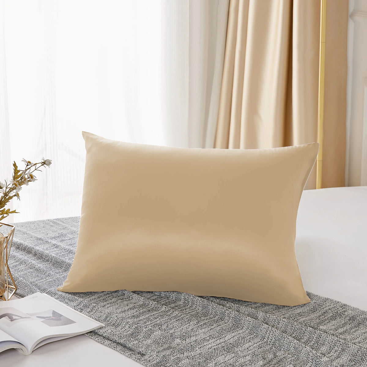A beige silk pillowcase resting on a gray linen fabric next to a window with sheer curtains.