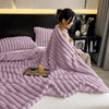 A fluffy lavender fleece throw blanket draped over a bed, next to pillows and a dark nightstand.
