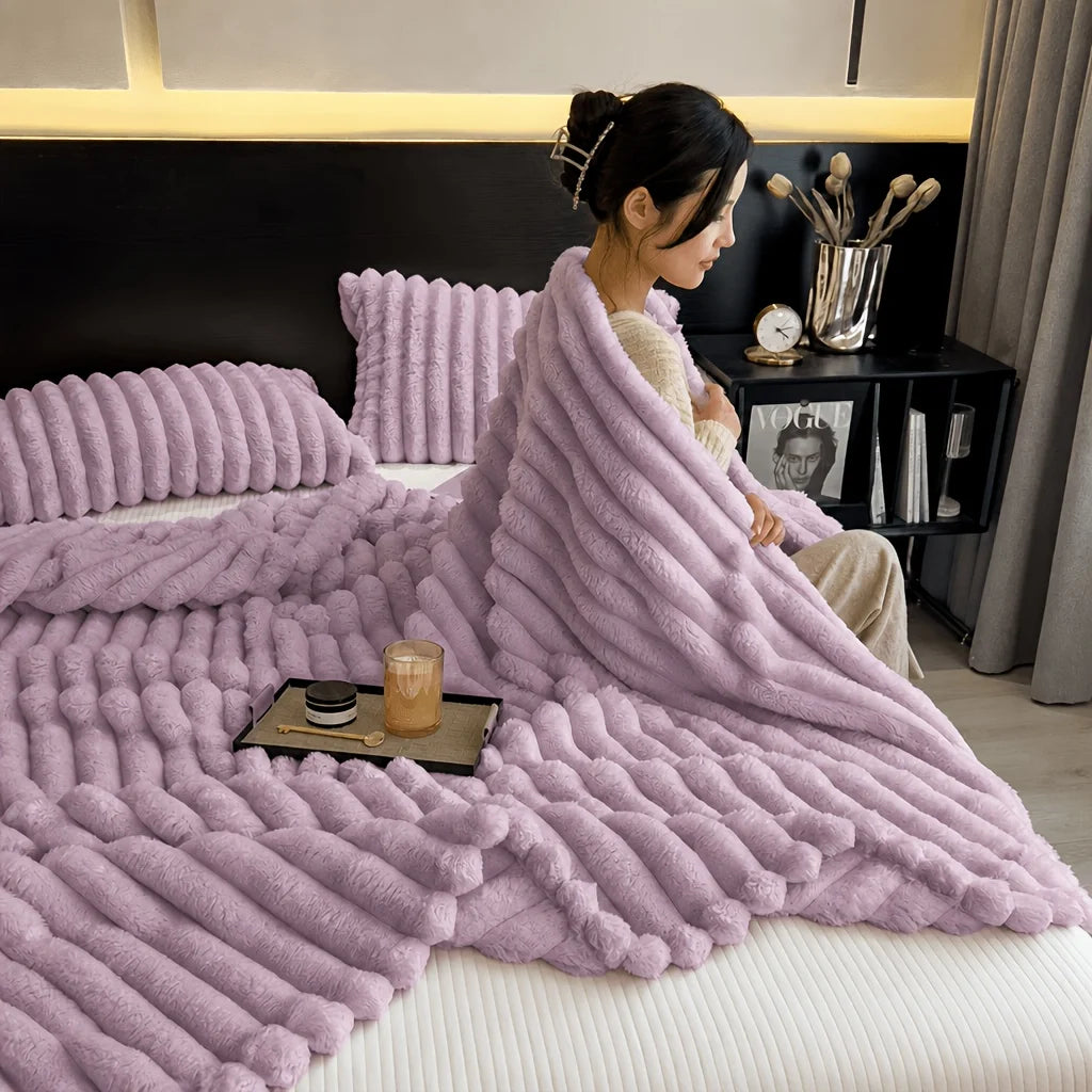 A fluffy lavender fleece throw blanket draped over a bed, next to pillows and a dark nightstand.