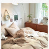 A bed dressed with beige and cream bedding, pillows, and a quilt set in a bright bedroom.