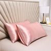 A set of pink satin silk pillows resting against a beige upholstered headboard and next to a lamp.